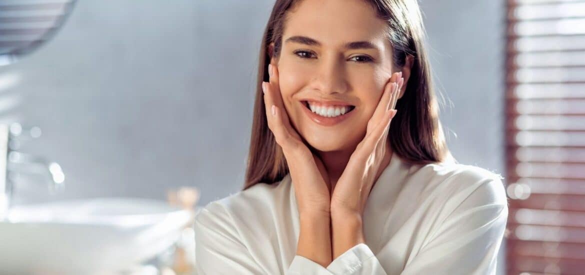 A beautiful young woman with a radiant, white smile gently touches her cheeks
